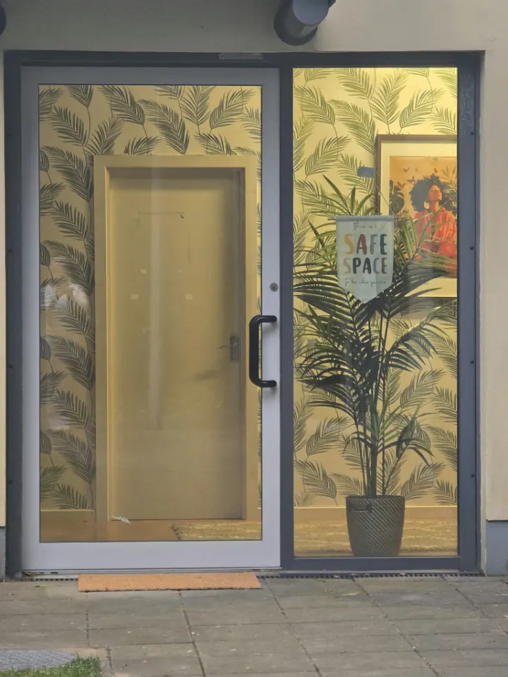 The glass front door of the counselling space, with palm-leaf wallpaper and a 'This is a safe space' sign inside